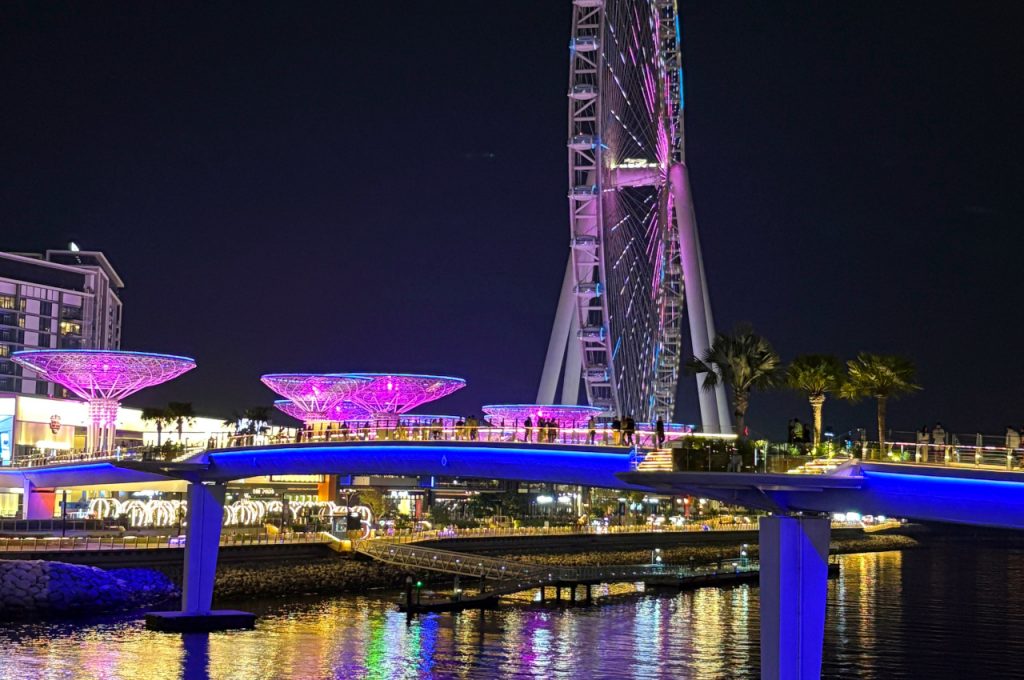 Night-time scene of Ain Dubai glowing in purple light, with a pedestrian bridge and waterfront lights reflecting on the sea.