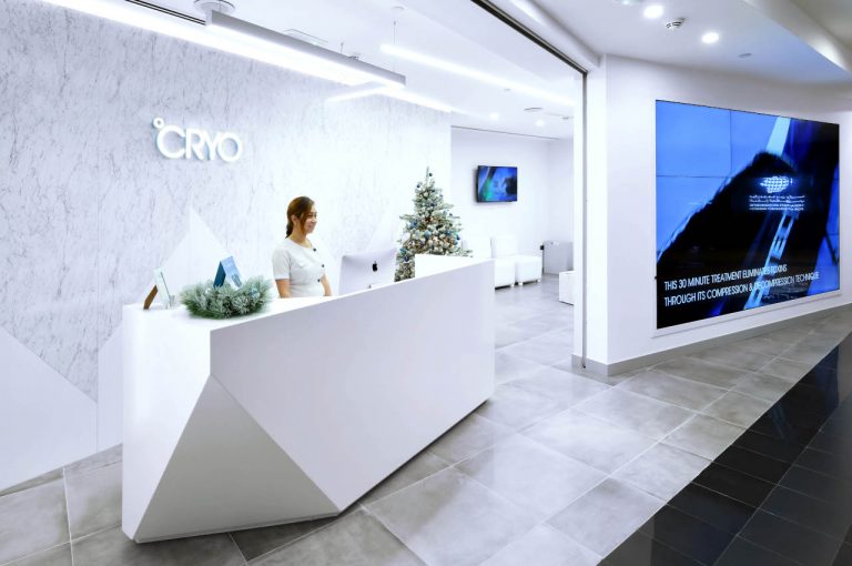 Small consultation room in a °CRYO centre with two beige chairs, a white desk, product shelf, posters on the wall and soft lighting.