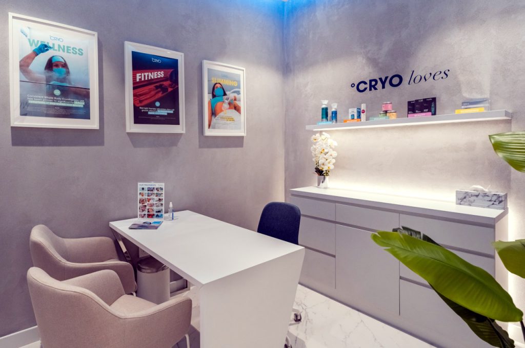 Modern °CRYO reception with a white geometric front desk, a staff member behind the counter, marble-style walls and a large digital screen in the hallway.