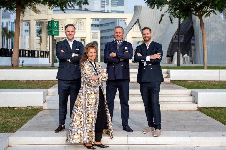 Four InvestMenter team members, three men in dark suits and one woman in a patterned long coat, standing with arms crossed on outdoor steps in Dubai Internet City.