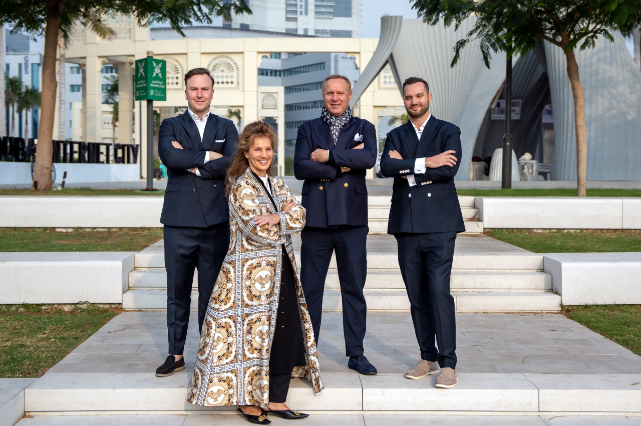 Four InvestMenter team members, three men in dark suits and one woman in a patterned long coat, standing with arms crossed on outdoor steps in Dubai Internet City.