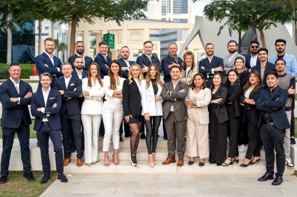 Large group photo of the InvestMenter team in business attire standing on steps in Dubai Internet City, with trees and office buildings in the background.
