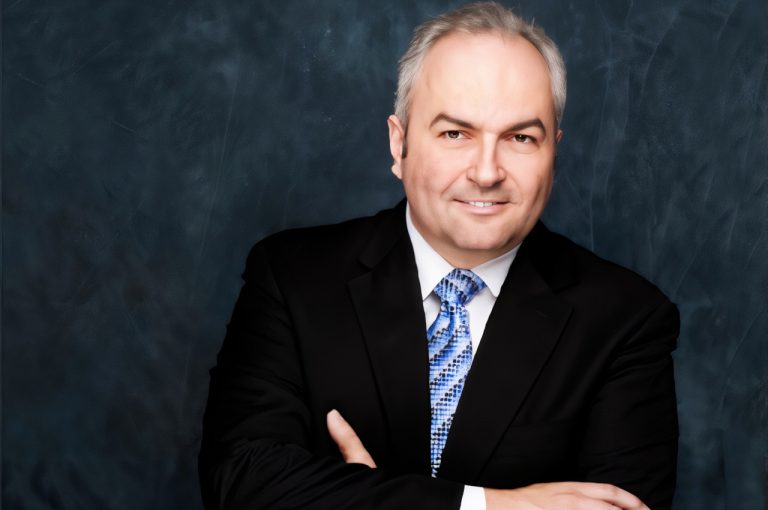 Formal studio portrait of Dr. Dany Kayle in a black suit and patterned blue tie, standing with arms crossed against a dark textured background.