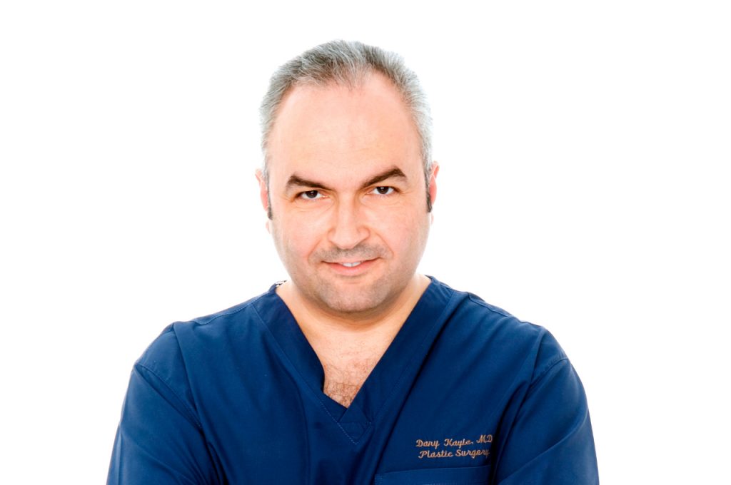 Dr. Dany Kayle wearing dark blue surgical scrubs, standing against a white background with arms folded and a focused expression.