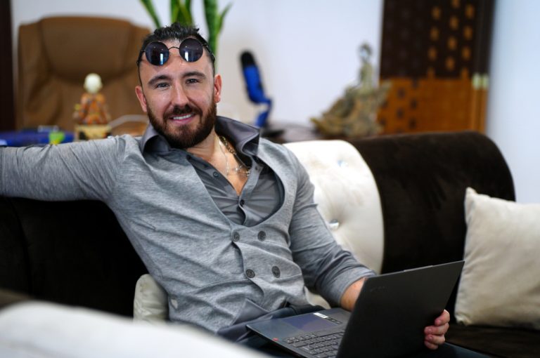 Mathieu Schmidt sitting relaxed on a sofa with a laptop on his lap, wearing a grey shirt and cardigan, looking at the camera in a casual office setting.
