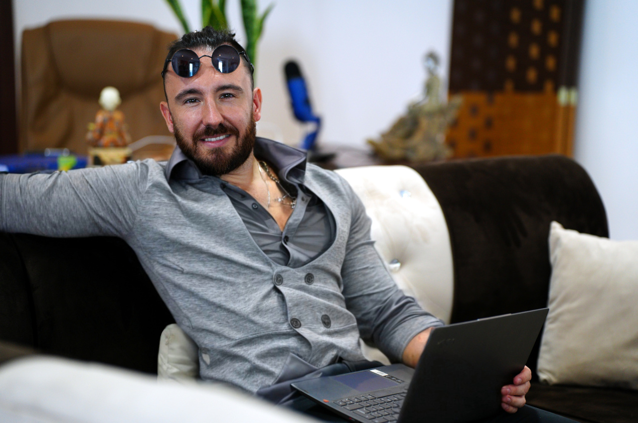 Mathieu Schmidt sitting relaxed on a sofa with a laptop on his lap, wearing a grey shirt and cardigan, looking at the camera in a casual office setting.