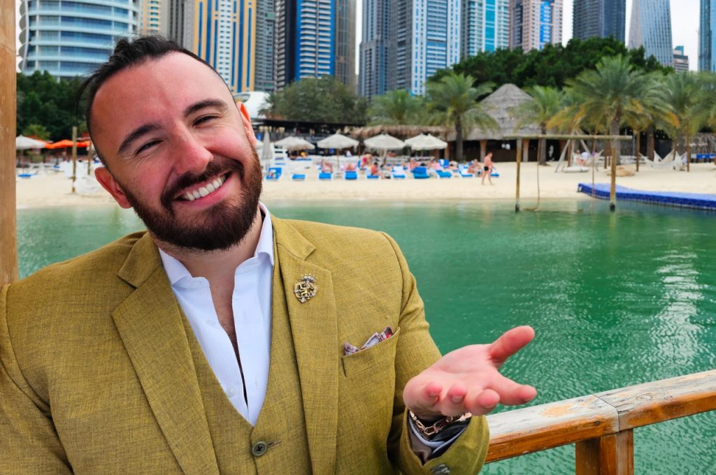 Portrait of Mathieu Schmidt in a mustard-coloured suit jacket, smiling at the camera on a waterfront terrace in Dubai with a beach and skyscrapers in the background.