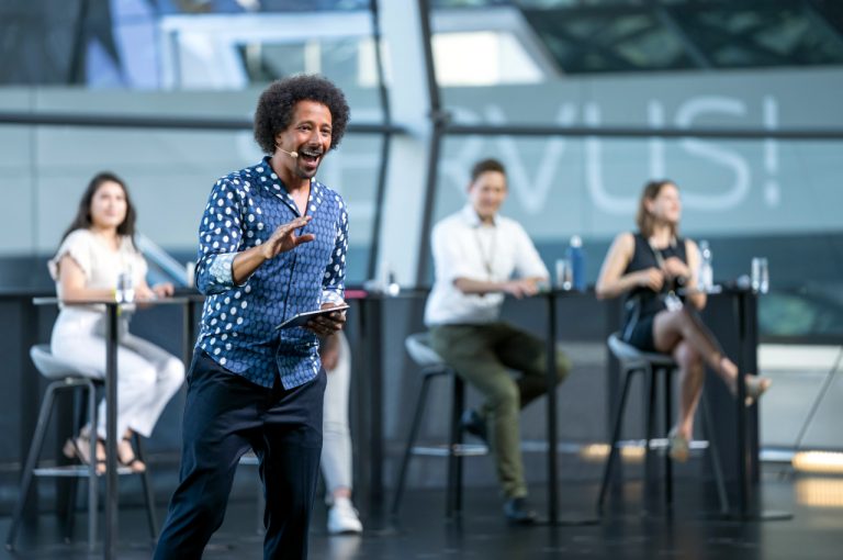 Flo Akinbiyi with curly hair in a polka dot shirt speaking on stage with a tablet in his hand, while three panelists sit at high tables in the background.
