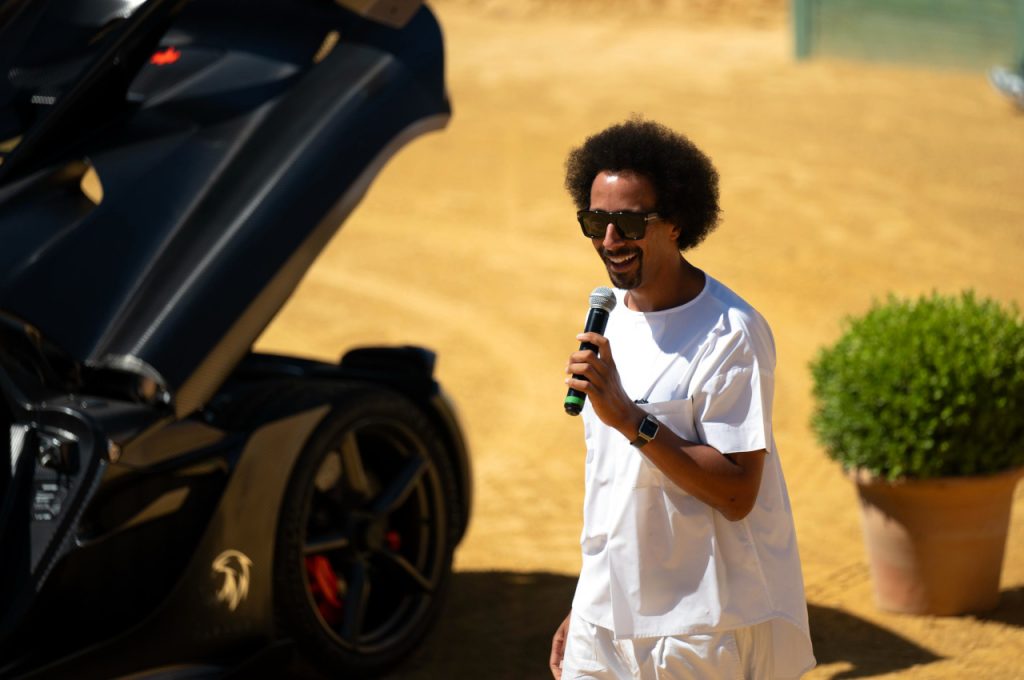 Flo Akinbiyi in a white outfit and sunglasses speaking into a handheld microphone next to a black supercar on a sunlit sand-coloured surface.
