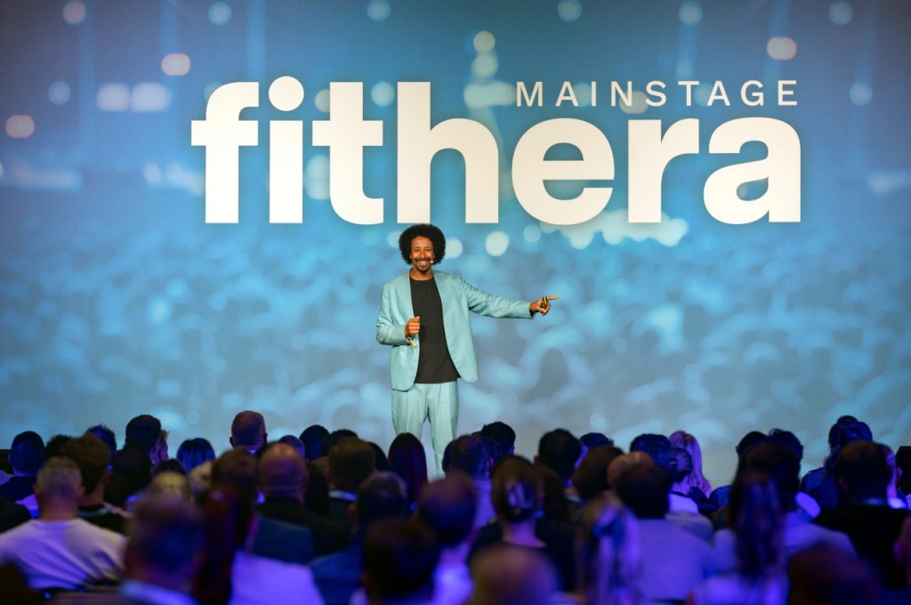 Flo Akinbiyi in a light-coloured suit standing in front of a huge screen reading “fithera mainstage”, addressing a seated audience in a darkened hall.