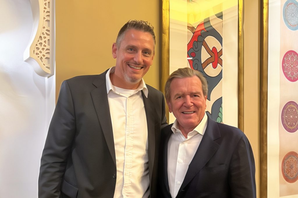 Mitch Bittermann in a dark suit standing beside Gerhard Schröder, both smiling in front of a decorative golden wall at an indoor event.