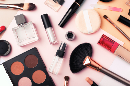 Flat lay of assorted beauty and makeup products, including brushes, perfume, lipstick, powder, lip gloss, and an eyeshadow palette, arranged on a soft pink background.