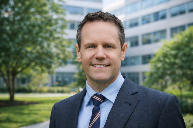 Portrait of Simon Lacey, a trade policy and geopolitical risk analyst, wearing a dark suit and striped tie outdoors.
