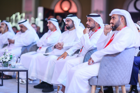 A group of men wearing traditional white Emirati attire seated in the front row at a formal conference, listening attentively to a speaker or presentation.