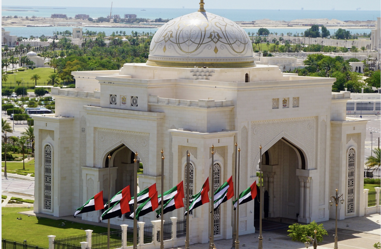 A striking view of a white palace surrounded by UAE flags and lush greenery under a clear sky.