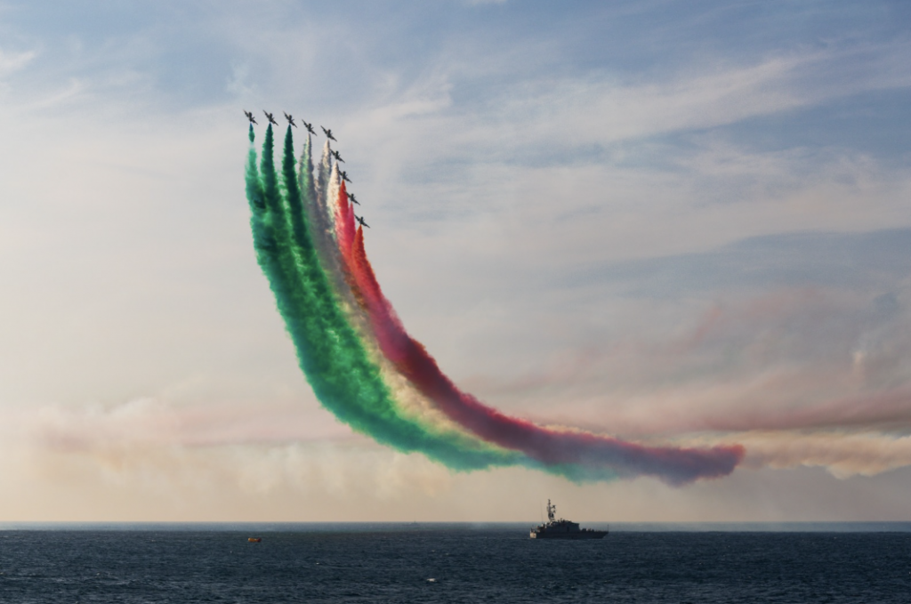 Aerobatic jets fly in formation above the sea, leaving sweeping smoke trails in the colors of the UAE flag—green, white, red, and black.